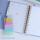 Open planner with gold spiral binding and a small sheet of colorful stickers on a white surface.